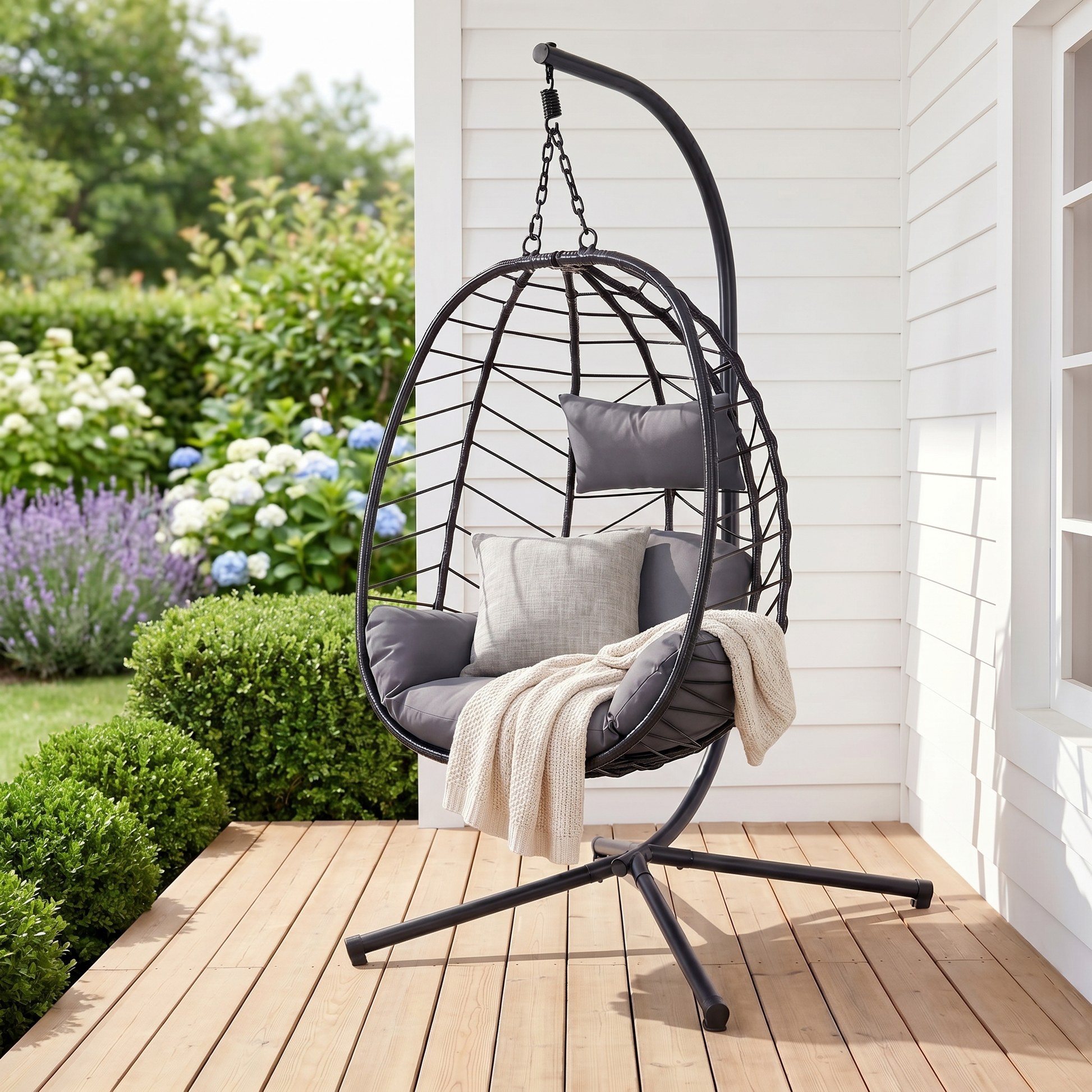 Black rattan egg swing chair with grey cushion on a porch in a temperate garden, square